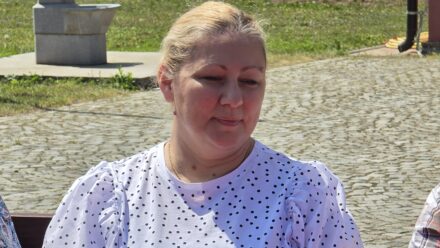 Žena sedi na klupi u parku, osunčana i nasmejana, u pozadini zelena trava i kamenje.