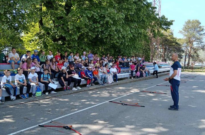 Grupa dece i odraslih na edukativnom predavanju na otvorenom, ispred sportskog terena.