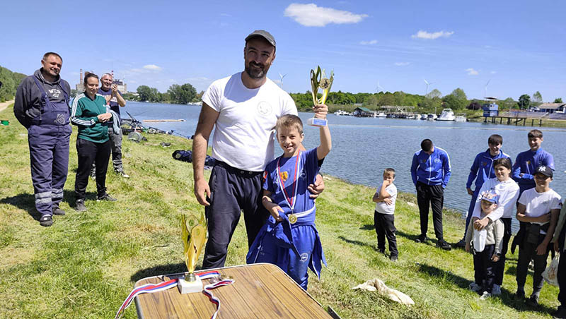 Otac i sin na takmičenju u sportu pored reke, sa medaljama i peharima.
