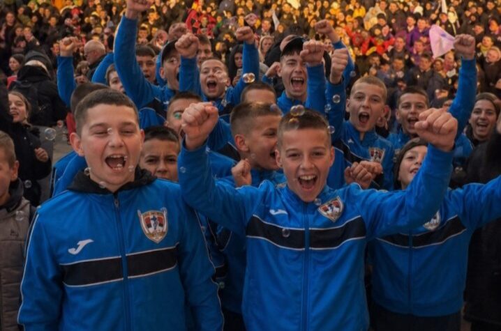 Vesela fudbalska ekipa dece u plavim dresovima sa grbovima, uz osmehe i podignute pesnice, na stadio.
