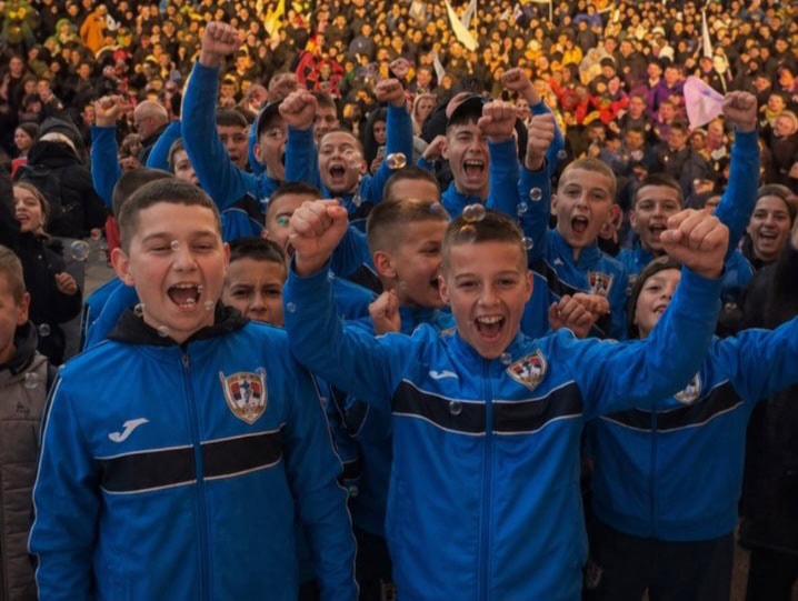 Vesela fudbalska ekipa dece u plavim dresovima sa grbovima, uz osmehe i podignute pesnice, na stadio.