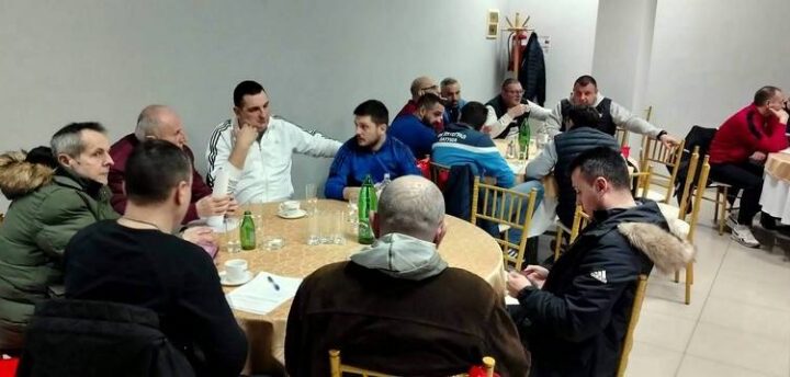 Sličica prikazuje grupu ljudi kako sede za stolom tokom sastanka ili diskusije u zatvorenom prostoru.