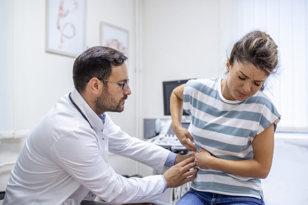 Patient telling physician about her pain and health problems during visit to hospital. Young woman complaining about back or kidney ache while sitting on examination bed at the doctor's office